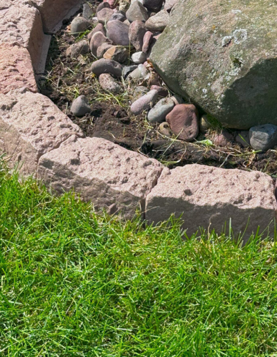 Row of light pink rectangular stones forming a border around a small rock garden bed with dirt and pebbles; green lawn in the foreground.