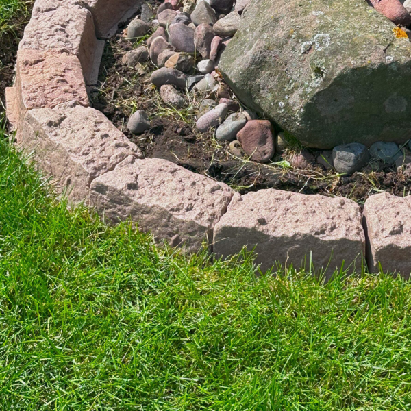 Row of light pink rectangular stones forming a border around a small rock garden bed with dirt and pebbles; green lawn in the foreground.
