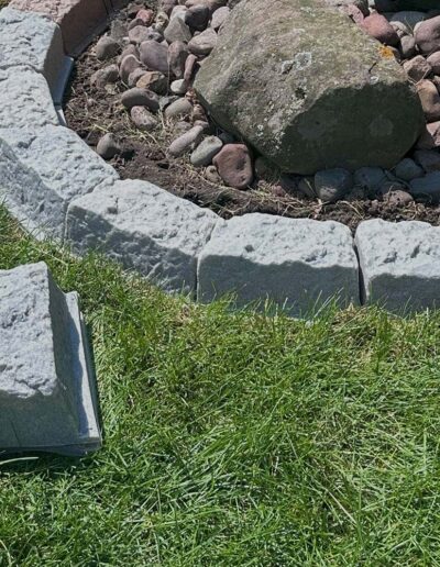 Gray curved stone edging forming a border between grass and a rock-filled garden bed with larger boulders nearby