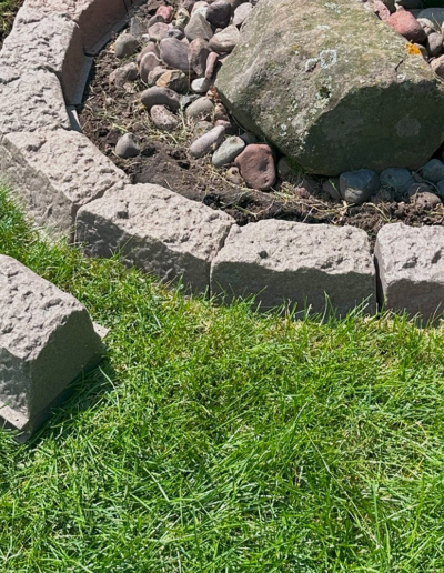 Curved border of grey stones forming a circular garden bed with a large mossy rock and smaller river rocks; green lawn in foreground.